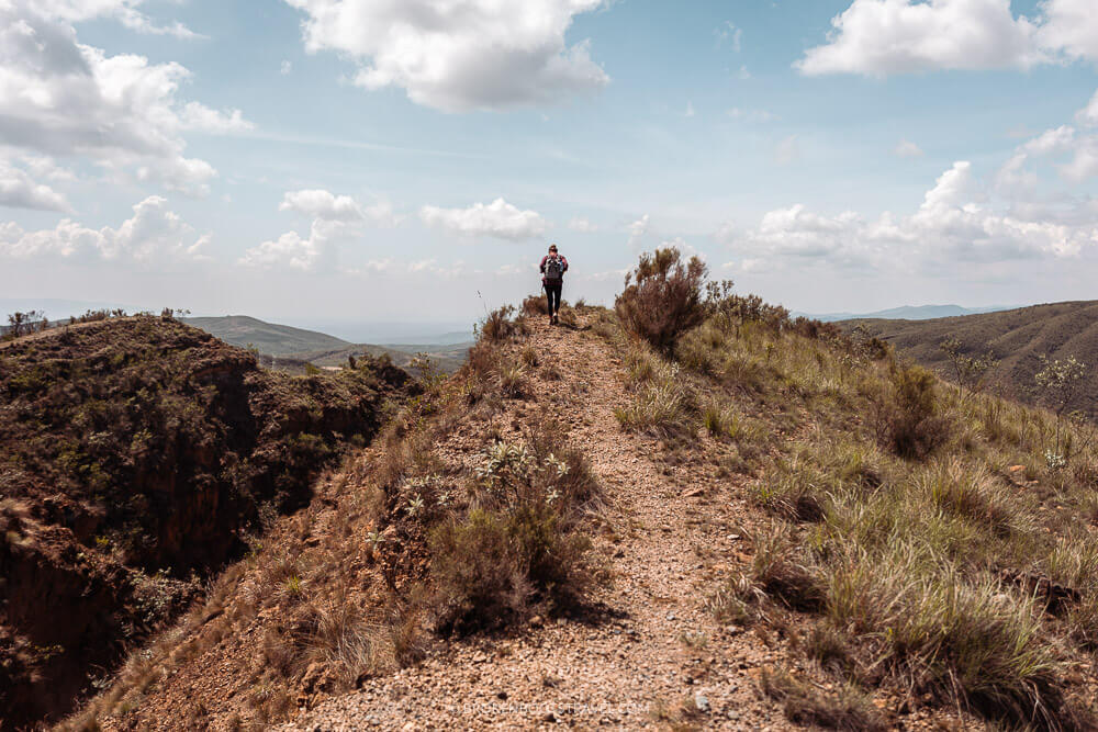 Girl wearing backpack walking alongside a crater in front of green mountains