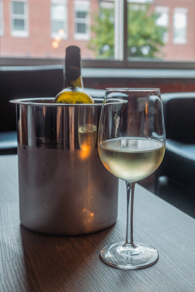 A glass of white wine and the bottle in front of a window