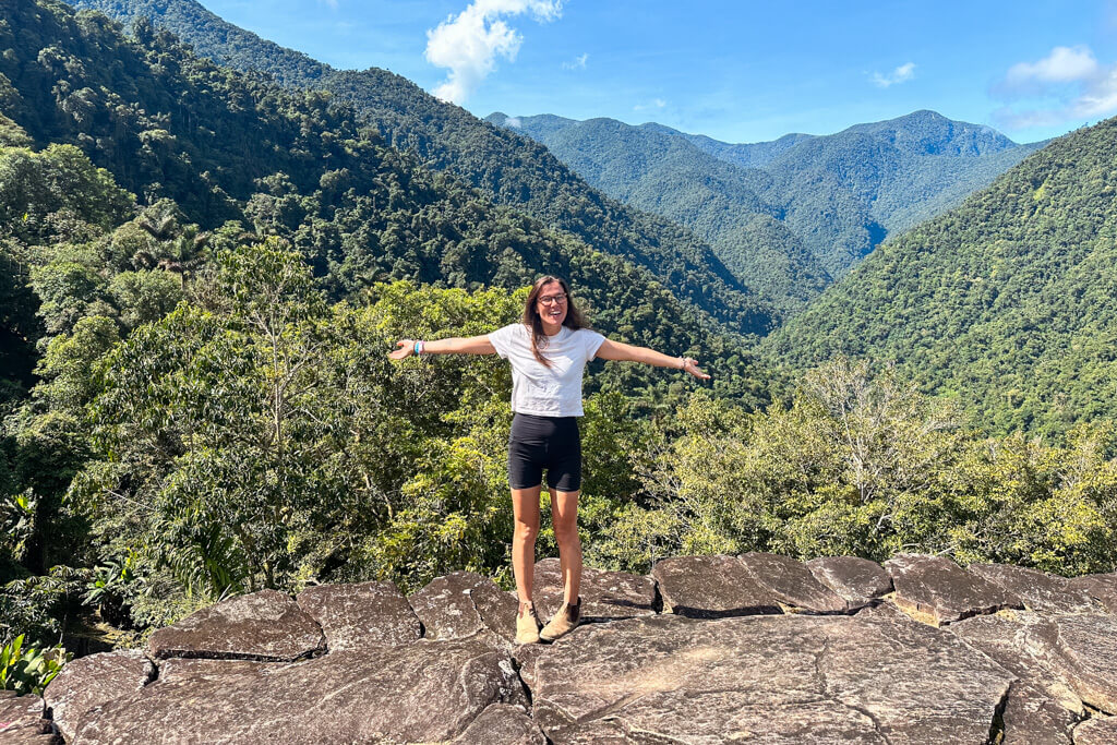 Girl in white shirt and black shorts stands in front of green mountains