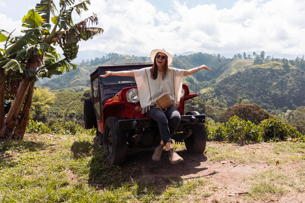 Girl in white poncho and white hat sits on a red Jeep in front of green mountains