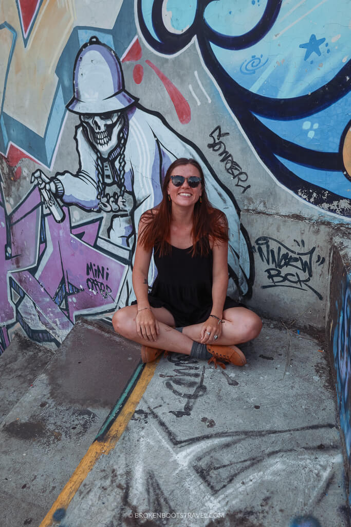 A girl in a black dress and sunglasses sits cross legged in front of a colorful mural