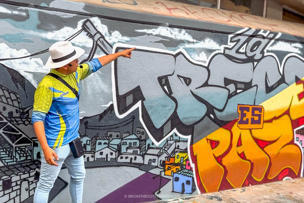 A guide in a blue and green shirt points to a mural that reads "la trece es paz"
