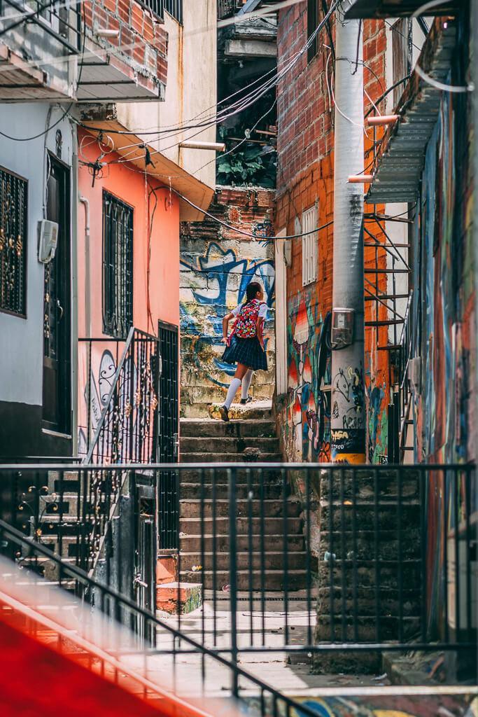 A girl in a school uniform and carrying a backpack walks through a neighborhood with murals. Comuna 13, Medellin, Colombia