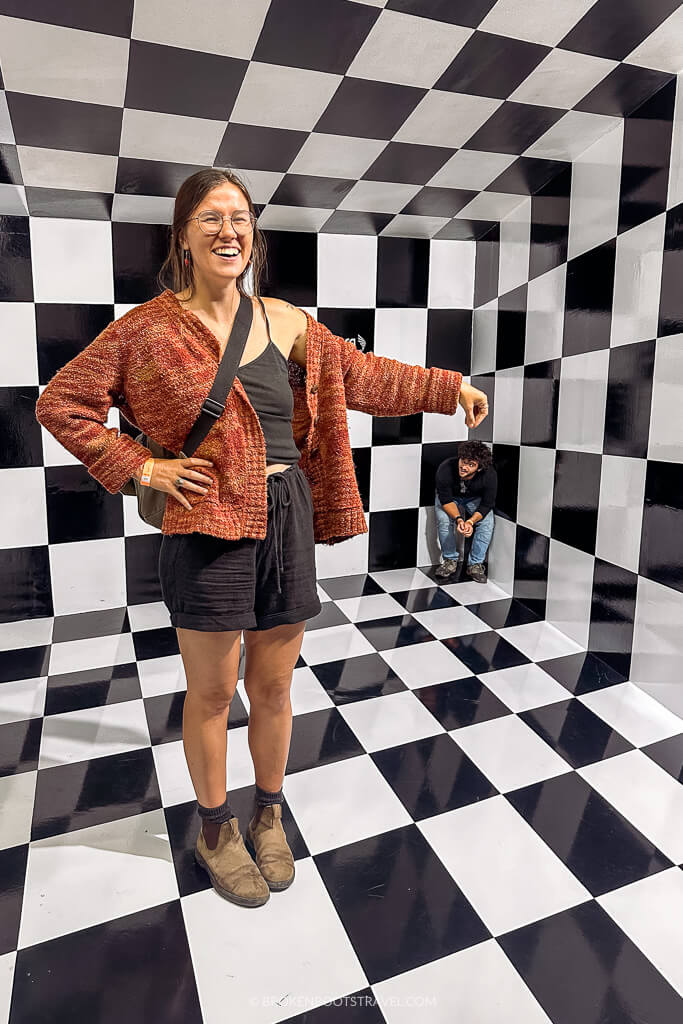 Girl in red sweater smiling in a checkerboard illusion room with man in black shirt in the corner