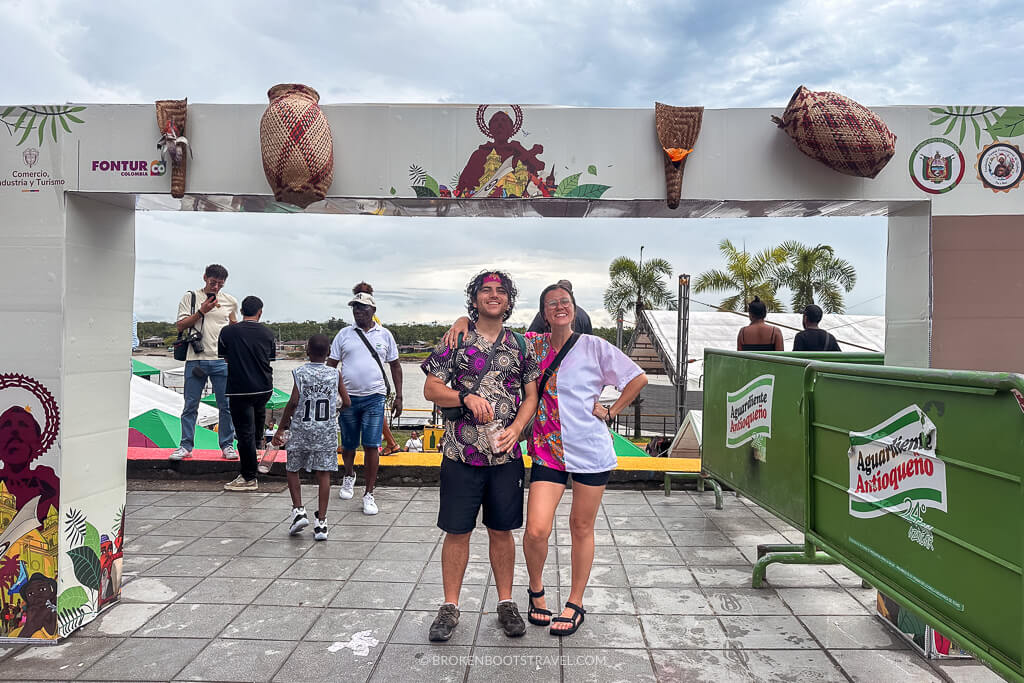 Two people wearing African wax print shirts in front of a sign for the San Pacho Festival